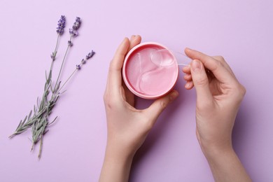 Photo of Woman taking under eye patch with spatula out of jar near lavender flowers on lilac background, top view. Cosmetic product