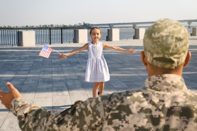 Little girl with flag of USA running towards her father in military uniform outdoors. Family reunion