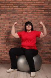 Happy overweight mature woman sitting on fitness ball near brick wall indoors