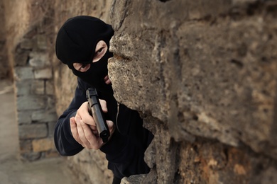 Masked man with gun hiding behind stone wall outdoors. Criminal offence