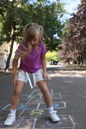 Photo of Little girl playing hopscotch drawn with chalk on asphalt outdoors. Happy childhood