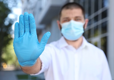 Man in protective face mask showing stop gesture outdoors, focus on hand. Prevent spreading of coronavirus