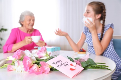 Preteen girl with her granny drinking tea at home, focus on flowers and card. Happy Mother's Day