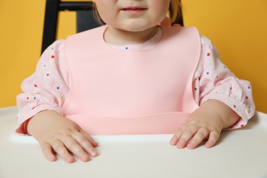Cute little baby wearing bib in highchair on yellow background, closeup