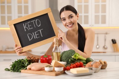 Happy woman holding chalkboard with words Keto Diet near different products in kitchen
