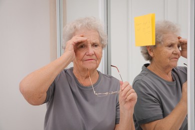 Forgetful senior woman near mirror with reminder note indoors. Age-related memory impairment