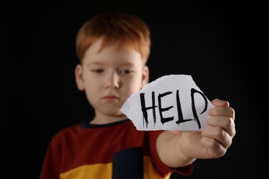 Photo of Little boy holding piece of paper with word Help against black background, focus on hand. Domestic violence concept