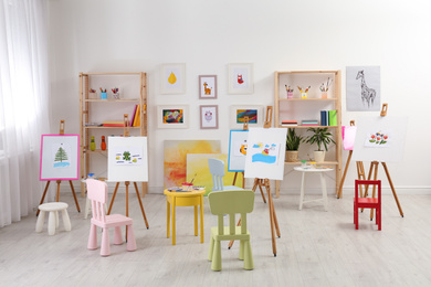 Easels with paintings and chairs for children in room