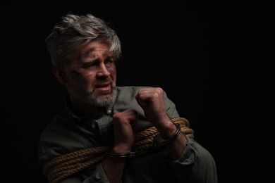 Photo of Frightened man tied with rope and handcuffs on black background, space for text. Hostage taking