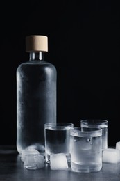 Bottle of vodka and shot glasses with ice on dark table against black background