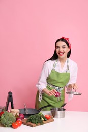 Young housewife cooking at white table on pink background. Space for text