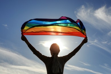 Woman holding bright LGBT flag against blue sky on sunny day, back view