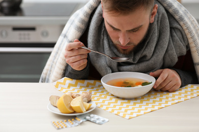 Sick young man eating tasty soup to cure flu at table in kitchen