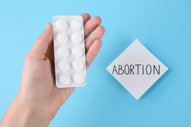 Woman holding pills near paper note with word Abortion on light blue background, top view