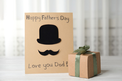 Greeting card with phrase HAPPY FATHER'S DAY I LOVE YOU DAD and gift box on white wooden table indoors