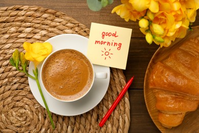 Cup of aromatic coffee with croissant, beautiful yellow freesias and Good Morning note on wooden table, flat lay