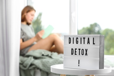Photo of Woman resting near window at home, focus on lightbox with phrase DIGITAL DETOX