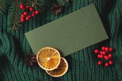 Flat lay composition with blank greeting card and Christmas decor on green knitted sweater. Space for text