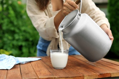 Photo of Woman pouring fresh milk from can into glass at wooden table outdoors, closeup