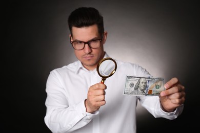 Photo of Expert authenticating 100 dollar banknote with magnifying glass against dark background, focus on hand. Fake money concept