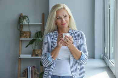 Photo of Beautiful woman with cup of hot drink near window at home
