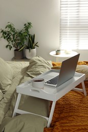 White tray with modern laptop and cup of aromatic tea on bed indoors