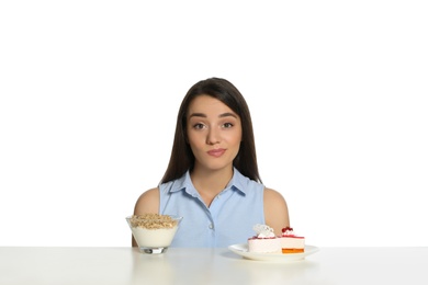 Doubtful woman choosing between yogurt with granola and cake at table on white background