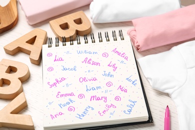 Notebook with different baby names and child's items on white wooden table, closeup