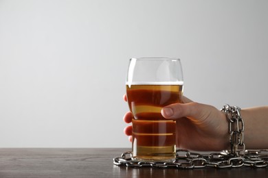 Woman with chained hand and glass of beer at wooden table against white background, closeup. Alcohol addiction