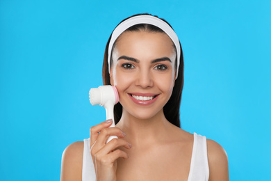 Young woman using facial cleansing brush on light blue background. Washing accessory