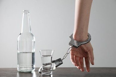 Woman in handcuffs with glass of vodka at wooden table against white background, closeup. Alcohol addiction