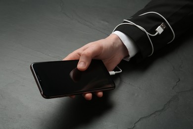 Internet addiction. Closeup of man holding phone at black table, hand tied to device with charging cable