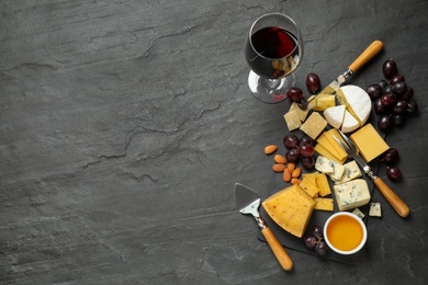 Cheese platter with specialized knives and fork and glass of red wine on grey table, flat lay. Space for text