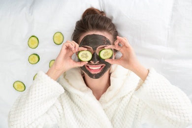 Young woman with facial mask lying and cucumber slices on bed, top view