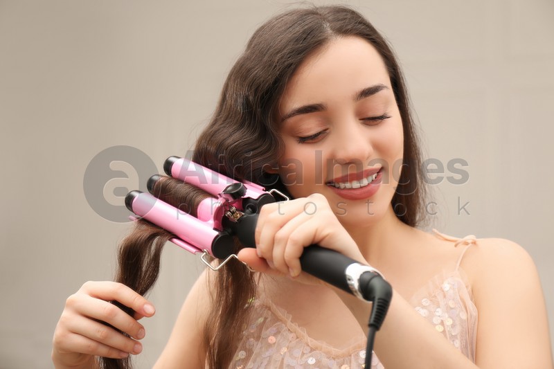 Young woman using modern curling iron indoors