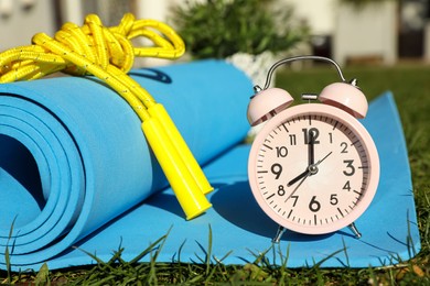 Photo of Alarm clock, fitness mat and skipping rope on green grass outdoors. Morning exercise