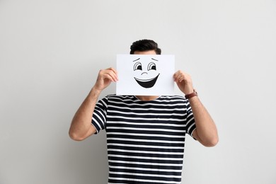 Man hiding emotions using card with drawn smiling face on white background