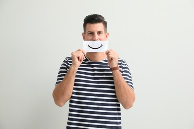 Man hiding emotions using card with drawn smile on white background