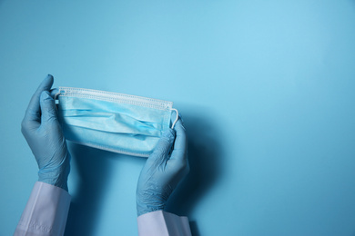 Doctor in latex gloves holding disposable face mask on light blue background, closeup with space for text. Protective measures during coronavirus quarantine