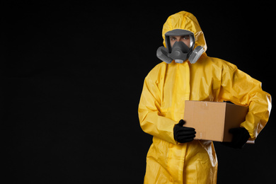 Man wearing chemical protective suit with cardboard box on black background, space for text. Prevention of virus spread