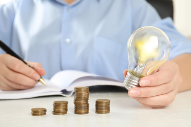 Woman with light bulb, notebook and coins at white table, closeup. Power saving