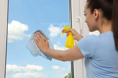 Photo of Young woman cleaning window glass with rag and spray indoors, closeup