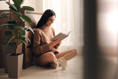 Photo of Woman with cup of drink and book sitting on warm floor at home. Heating system