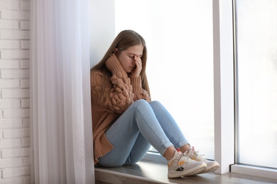 Upset teenage girl sitting alone near window indoors