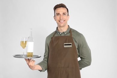 Handsome waiter holding tray with glass and bottle of wine on light background