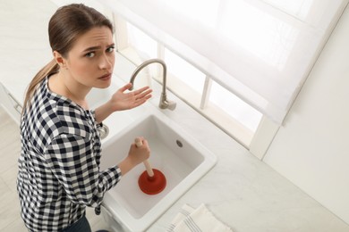 Upset young woman using plunger to unclog sink drain in kitchen, above view