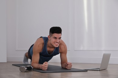 Handsome man doing plank exercise on yoga mat while watching online class indoors
