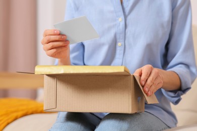 Photo of Woman holding blank greeting card near package with gift indoors, closeup