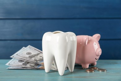 Ceramic model of tooth, piggy bank and money on light blue wooden table. Expensive treatment