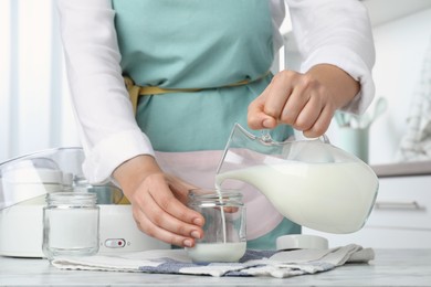 Woman pouring milk into glass jar at white marble table, closeup. Making yogurt